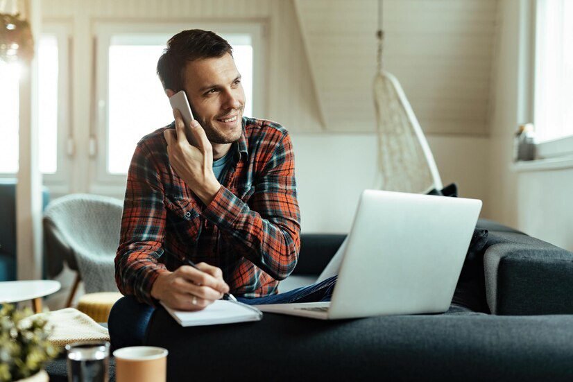 jovem feliz tomando notas e se comunicando no celular enquanto usa um computador na sala de estar